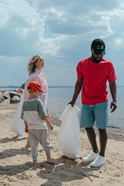 A group of three people, including an adult male and two children, are on a sandy beach near a body of water during daytime, participating in a rubbish removal activity. The man, dressed in a red T-shirt, light blue shorts, and a black cap, holds a white plastic bag in his right hand, appearing to collect litter from the sand. One of the children, wearing a red cap with a pattern, a light-colored top, and grey pants, stands close to him, observing or waiting to help. The other child, a girl with blonde curly hair, is dressed in a light pink jacket and light trousers, standing to the left and looking towards the water, possibly holding or about to place rubbish into the bag. The background features a cloudy sky and a calm waterfront with rocks along the shoreline, indicating a peaceful outdoor environment suitable for private waste collection or environmental cleanup activities, reminiscent of on-site rubbish removal services facilitated by companies like Rubbish Removal Enfield, focused on eco-friendly and responsible waste handling practices.