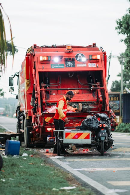 A waste collection worker wearing a high-visibility orange vest and dark trousers is operating a large red rubbish compactor truck on a roadside, with the truck’s rear open, revealing the internal compaction mechanism. The worker is standing next to a small metal enclosure attached to a motorbike, which holds a large black plastic garbage bag. The scene is set on a paved urban street with a grassy verge on the left, and a blue container partially visible nearby. In the background, there are trees, utility poles, and signage, with overcast skies overhead. The truck is spacious and designed for on-site rubbish collection, with visible warning lights and reflective markings. This scene exemplifies an independent waste collection service, aligning with the context of alternative rubbish removal methods supported by companies like Rubbish Removal Enfield, serving non-local authority waste disposal needs. The image captures a typical urban waste collection operation, illustrating the process of private waste handling and rubbish removal in a professional environment.