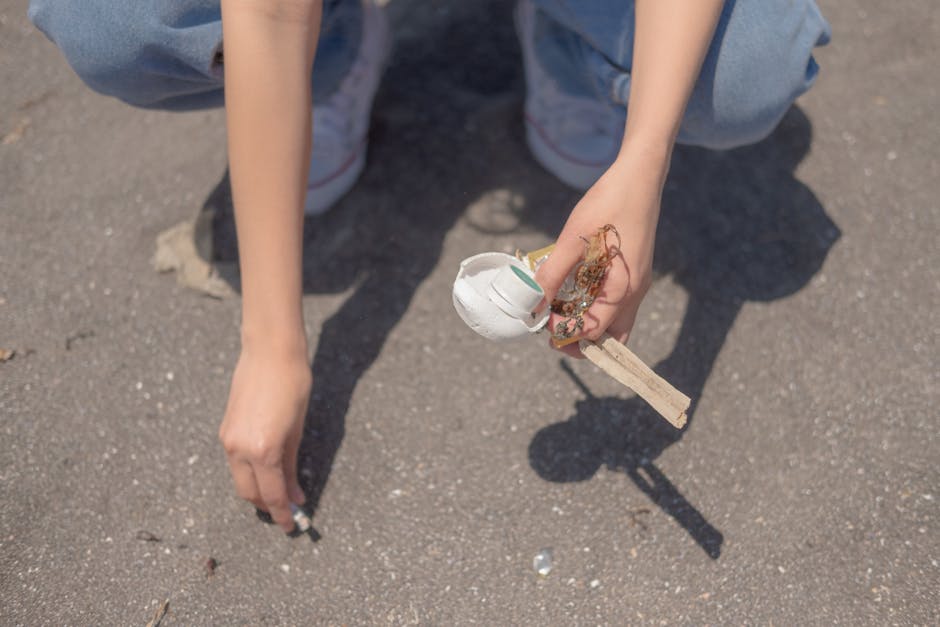 A person crouching on a paved surface, holding a small white plastic container with torn and frayed edges, attached to a wooden stick or handle, with visible dirt and debris around. The individual is wearing light blue jeans and white sneakers with red accents, and their other hand is reaching towards the ground. The background shows a rough asphalt area with small stones and dust, highlighting the act of littering or waste disposal outdoors. The scene emphasizes the importance of proper rubbish management and can relate to private waste collection services like those offered by Rubbish Removal Enfield, illustrating the need for responsible waste handling and cleanup.