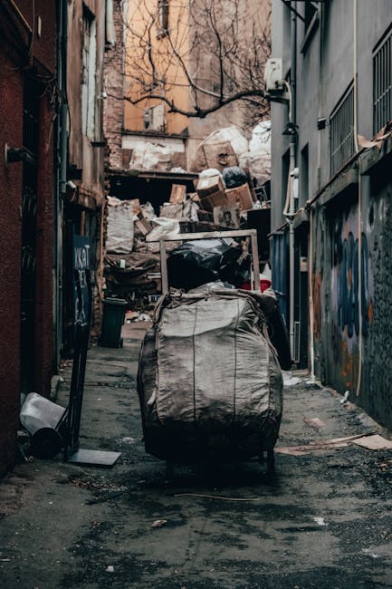 This image depicts a narrow urban alleyway cluttered with overflowing piles of mixed waste, primarily consisting of large black and gray plastic rubbish bags, some torn open revealing debris inside. The bags are stacked against a backdrop of weathered brick and concrete walls covered in graffiti and peeling paint. A large, wheeled laundry or waste trolley with a sturdy metal frame and fabric cover stands prominently in the foreground, surrounded by scattered smaller items, including a plastic container and a metal rod. Behind the trolley, a dense accumulation of discarded cardboard boxes, paper, and various unidentifiable rubbish creates a chaotic scene. The environment appears damp, with darkened patches on the pavement, and faint natural light filters through leafless trees and dilapidated structures, suggesting an outdoor setting in an alleyway between buildings. This scene exemplifies improper waste disposal in urban areas where private rubbish collection services such as Rubbish Removal Enfield might be employed as an alternative to conventional council services for efficient on-site clearance and waste management.