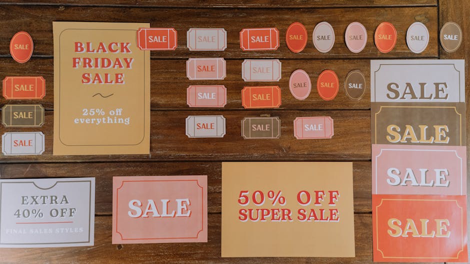 A collection of various promotional sale signs and labels displayed on a wooden surface, including rectangular, oval, and circular shapes in shades of red, white, pink, and beige. Prominent signs advertise discounts such as 'Black Friday Sale' with 25% off everything, '50% Off Super Sale', and 'Extra 40% Off Final Sales Styles'. The signs feature bold typography, decorative borders, and contrasting colors to attract attention. The arrangement includes both small individual labels and larger posters, with some overlapping slightly, creating a cluttered but organized display typical for retail promotional displays. The wooden background provides a warm, textured surface that contrasts with the bright and pastel-colored signs, emphasizing their visibility. This scene appears to be part of a sales display for a store or market, showcasing different promotional offers designed to attract customers. The overall setup suggests a focus on reducing stock or promoting seasonal discounts, aligning with practices around private, independent sales, or clearance events often associated with waste reduction in retail environments, indirectly supporting reuse and repurposing efforts. The lighting is even and natural, enhancing textual clarity and detail, making all signs easily readable for accessibility purposes.