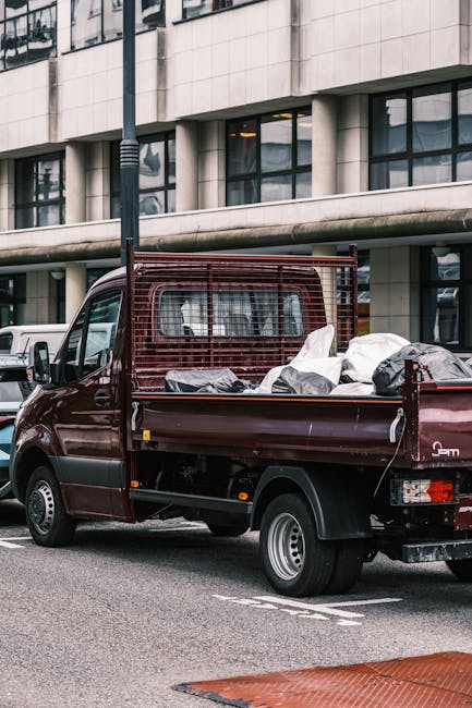 A small, dark red flatbed truck parked on a city street in front of a modern office building with large glass windows and a tiled facade. The truck's cargo area is filled with various bags and loose rubbish, including white and black plastic sacks, some of which are partially open or crumpled. The vehicle features a protective red metal grid structure along the cab's rear window, and its rear lights and mudguards are visible. The scene is illuminated by natural daylight, with a streetlamp post nearby and several other parked vehicles in the background. This image exemplifies an independent rubbish collection vehicle used for on-site clearance and private waste handling, supporting alternative disposal methods outside of local authority services, and aligns with rubbish removal services like those provided by Rubbish Removal Enfield.