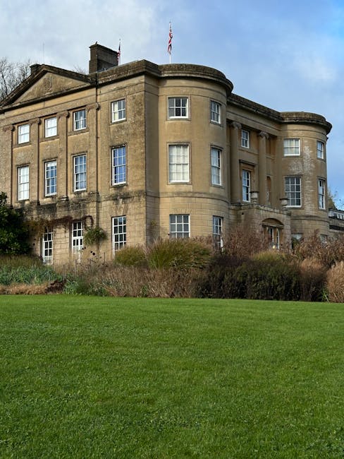 A historic, multi-story stone building featuring a rounded corner with rectangular windows arranged symmetrically, some with white framing. The facade has a weathered finish in light beige tones, with visible signs of age and minor weathering. The building's roofline displays a flat section with a flagpole flying the Union Jack, and a chimney protrudes from the roof. In the foreground, there is a well-maintained green lawn with neatly trimmed grass and some sparse low shrubs and bushes along the base of the building, suggesting a landscaped area surrounding the structure. The sky above is partly cloudy with patches of blue visible, providing natural lighting that highlights the building's textures and architectural details. The scene evokes an impression of an elegant, historic residence or institutional building, with no visible activity or people present, and the context suggests it may be part of a private estate or public grounds that require occasional waste clearance, which a company like Rubbish Removal Enfield might assist with when maintaining the site.