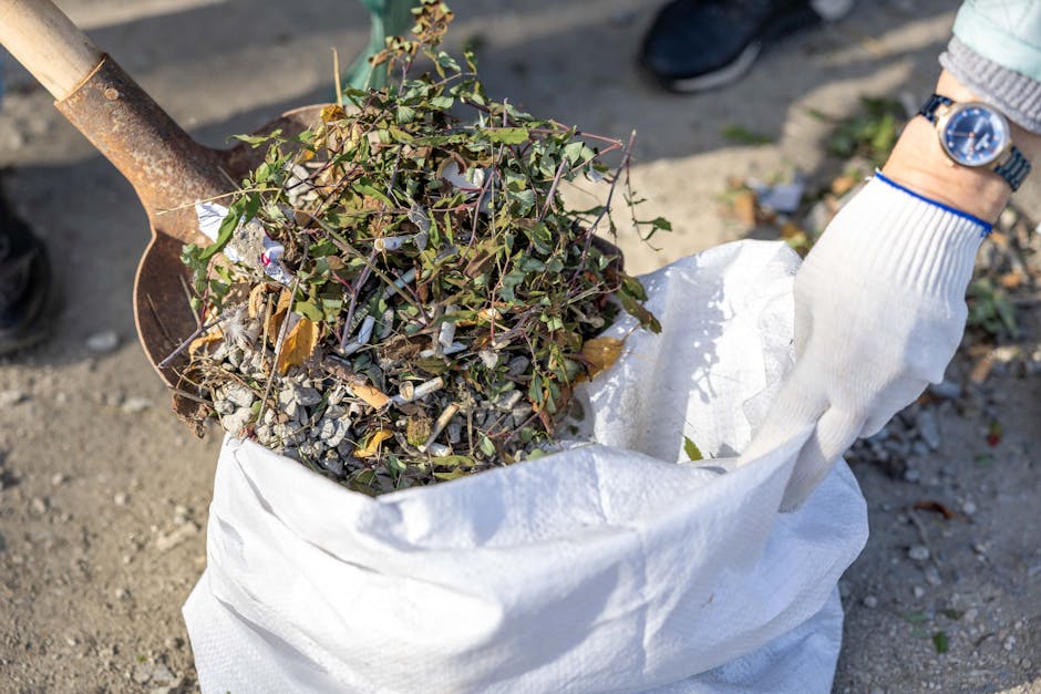 A person wearing a white glove and a wristwatch is holding a large white woven polypropylene sack filled with garden debris, including small branches, green leaves, and shredded plant material. The sack is situated outdoors on a gravel surface, with some dirt and small stones visible in the background. A rusty metal shovel with a wooden handle is partially inserted into the sack, suggesting that the debris is being collected for disposal or removal. The scene appears to be part of an outdoor waste clearance activity, supported by the presence of gardening or landscaping tools, consistent with private rubbish removal services offered by Rubbish Removal Enfield. The natural lighting highlights the textures of the leaves, twigs, and gravel, emphasizing the manual process of site clearance or garden waste removal.