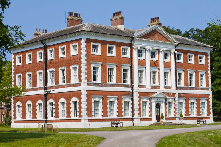 A historic, multi-story stone building featuring a rounded corner with rectangular windows arranged symmetrically, some with white framing. The facade has a weathered finish in light beige tones, with visible signs of age and minor weathering. The building's roofline displays a flat section with a flagpole flying the Union Jack, and a chimney protrudes from the roof. In the foreground, there is a well-maintained green lawn with neatly trimmed grass and some sparse low shrubs and bushes along the base of the building, suggesting a landscaped area surrounding the structure. The sky above is partly cloudy with patches of blue visible, providing natural lighting that highlights the building's textures and architectural details. The scene evokes an impression of an elegant, historic residence or institutional building, with no visible activity or people present, and the context suggests it may be part of a private estate or public grounds that require occasional waste clearance, which a company like Rubbish Removal Enfield might assist with when maintaining the site.