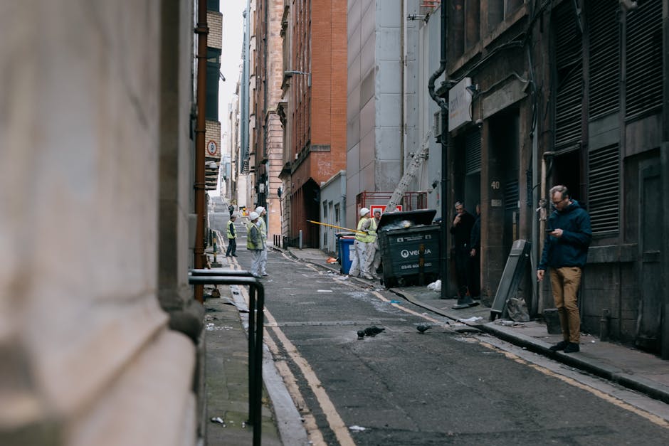 A narrow urban street scene showing a small amount of rubbish and debris along the asphalt pavement, with some paper and plastic scattered near the curbs. Several workers in high-visibility jackets and helmets are engaged in a waste collection or site clearance activity, standing around a large black skip bin positioned on the right side of the alleyway, partially open with waste visible inside. The surrounding buildings are tall, multi-storey structures with brick and stone facades, featuring windows and external pipes. One individual on the far right is wearing casual clothing and using a mobile phone, while others are focused on their task, with a few pigeons on the ground nearby. The environment appears to be an enclosed city courtyard or alley with limited natural light, creating a muted, subdued atmosphere suitable for private waste handling or independent rubbish removal services, such as those offered by Rubbish Removal Enfield, in support of alternative disposal options within urban areas.