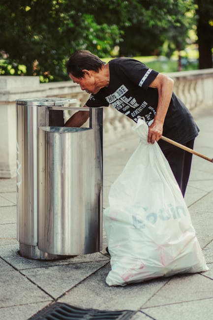 An elderly woman with short dark hair, dressed in a black T-shirt and dark trousers, is seen disposing of waste into a tall, cylindrical, stainless steel public rubbish bin outside on a paved pathway. She is holding a large white plastic bag filled with rubbish in her left hand while using her right hand to reach into the bin. The bin is positioned against a low stone wall with a decorative balustrade, set amid a green environment with trees and foliage in the background. The scene appears to be taking place during daytime under natural light, emphasizing a routine act of waste disposal. The stainless steel surface of the bin reflects some of the surrounding environment. This setting mirrors the typical outdoor spaces where private waste collection services, such as those offered by Rubbish Removal Enfield, facilitate alternative rubbish removal approaches outside of municipal systems.