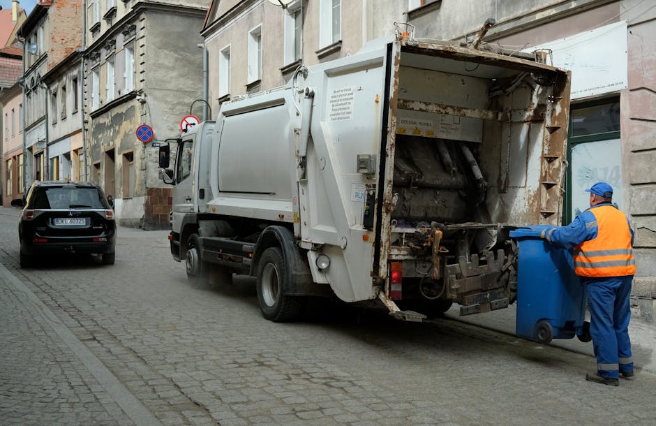 A street scene featuring a white refuse collection truck with an open rear compartment for waste loading parked on a cobblestone road in front of old multi-story buildings with weathered facades. A sanitation worker dressed in blue and orange high-visibility clothing and a matching cap is positioned on the right side, actively lifting a blue waste bin towards the truck’s rear hatch. The truck's surface shows signs of rust and grime, especially around the opening. To the left of the truck, a black vehicle is parked, and in the background, there are traffic signs indicating no parking zones. The environment appears quiet and residential, with natural daylight illuminating the scene, emphasizing the textures of the cobblestones, the worn building surfaces, and the metallic finish of the garbage truck. Rubbish removal services provided by Rubbish Removal Enfield are relevant here as part of alternative waste handling methods for urban household and commercial waste disposal, situated within the broader context of independent collection options outside the local authority’s regular schedule.