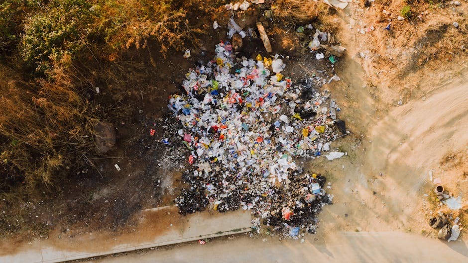 A large heap of mixed waste and landfill debris is gathered on a patch of bare, uneven earth near a concrete curb, with some evidence of charred or burnt materials at the base. The rubbish consists primarily of plastic bottles, food packaging, paper, and other household waste, with various colors including white, yellow, blue, black, and red. Surrounding the debris are sparse, dried bushes on one side and exposed soil with a reddish-brown tone. The scene appears to be an unmanaged or informal rubbish disposal area, possibly an unofficial dumping site, with the waste spread over a small section of ground adjacent to a paved pathway or street. The overall environment suggests an outdoor, outdoor urban or suburban setting where waste has been discarded, emphasizing the need for proper rubbish removal services such as those provided by Rubbish Removal Enfield, to facilitate controlled and environmentally responsible waste management and disposal.