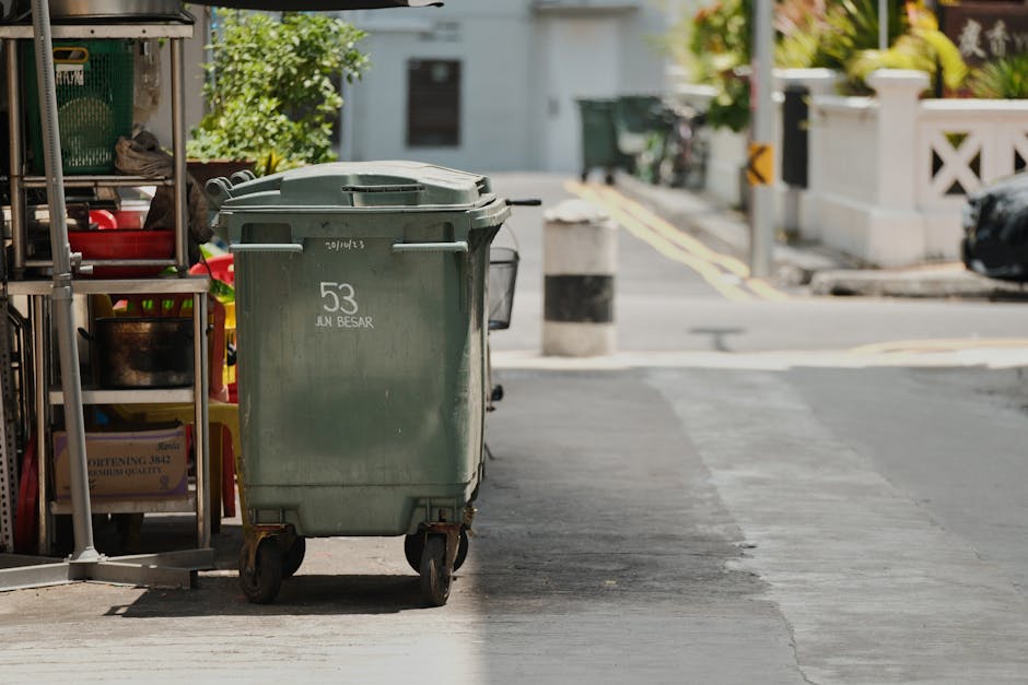 A street scene featuring a white refuse collection truck with an open rear compartment for waste loading parked on a cobblestone road in front of old multi-story buildings with weathered facades. A sanitation worker dressed in blue and orange high-visibility clothing and a matching cap is positioned on the right side, actively lifting a blue waste bin towards the truck’s rear hatch. The truck's surface shows signs of rust and grime, especially around the opening. To the left of the truck, a black vehicle is parked, and in the background, there are traffic signs indicating no parking zones. The environment appears quiet and residential, with natural daylight illuminating the scene, emphasizing the textures of the cobblestones, the worn building surfaces, and the metallic finish of the garbage truck. Rubbish removal services provided by Rubbish Removal Enfield are relevant here as part of alternative waste handling methods for urban household and commercial waste disposal, situated within the broader context of independent collection options outside the local authority’s regular schedule.